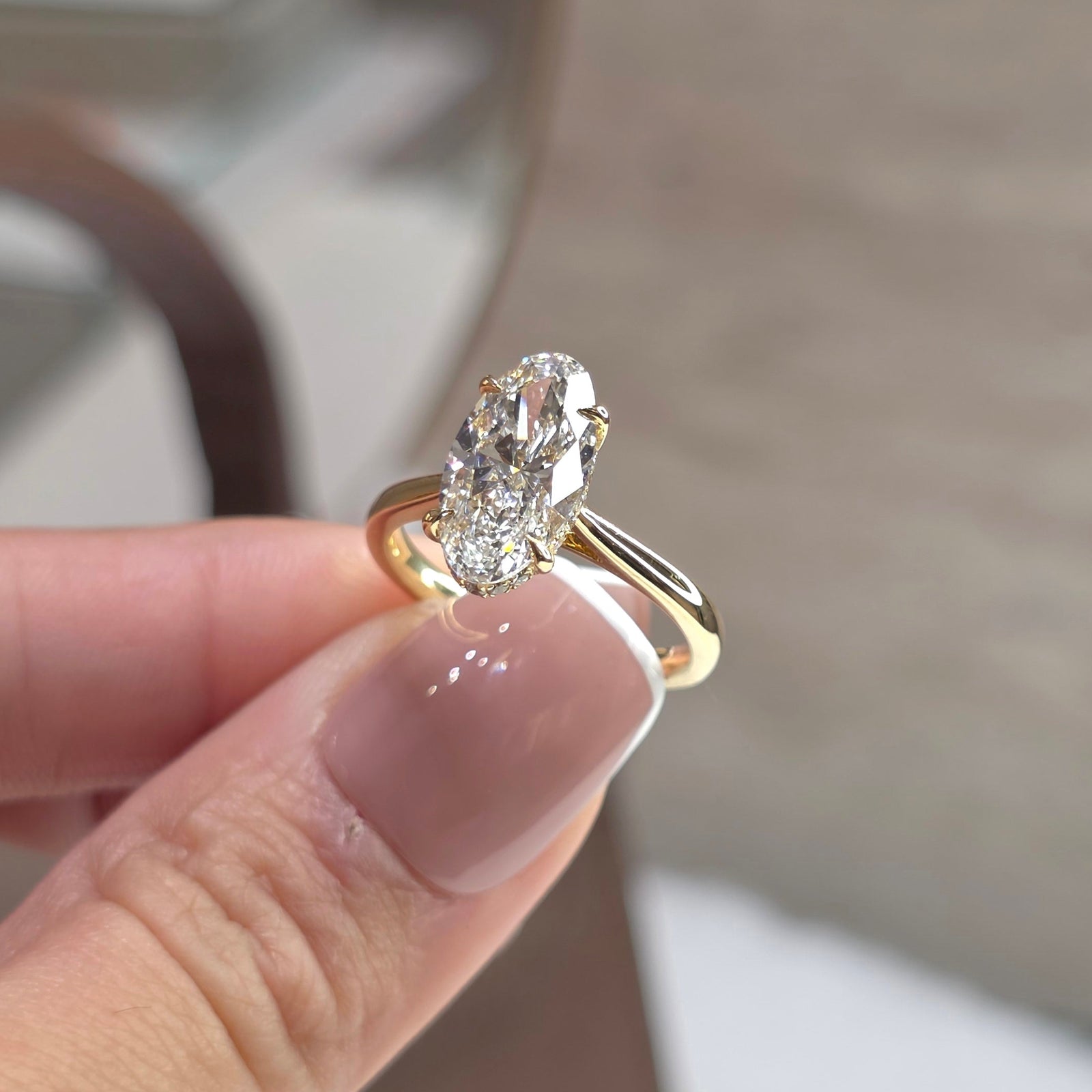 A gold ring with an oval-cut clear gemstone held between fingers. The ring features a yellow gold band with a split shank design and a bezel-set oval stone.