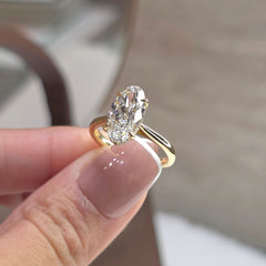 A gold ring with an oval-cut clear gemstone held between fingers. The ring features a yellow gold band with a split shank design and a bezel-set oval stone.