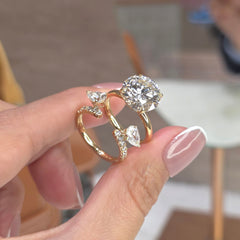 A gold ring with a large round-cut clear center stone and three smaller heart-shaped clear stones, held between fingers. The ring features pavé-set accent stones along the band.
