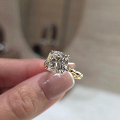 A close-up of a hand holding a yellow gold ring with a cushion-cut clear gemstone, set against a softly blurred beige background.