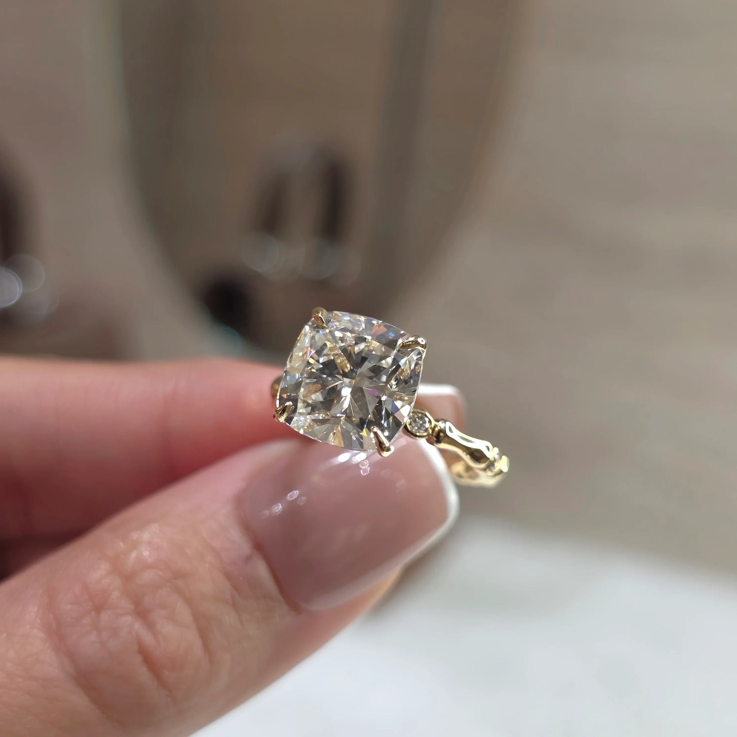 A close-up of a hand holding a yellow gold ring with a cushion-cut clear gemstone, set against a softly blurred beige background.