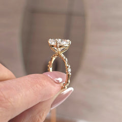 A close-up of a gold ring with a large clear rectangular gemstone and small side stones, held between fingers against a softly blurred beige background.