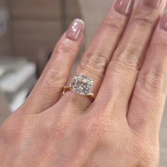 A close-up of a hand wearing a gold ring with a cushion-cut clear gemstone. The ring is shown on a finger with manicured nails, set against a softly blurred indoor background.