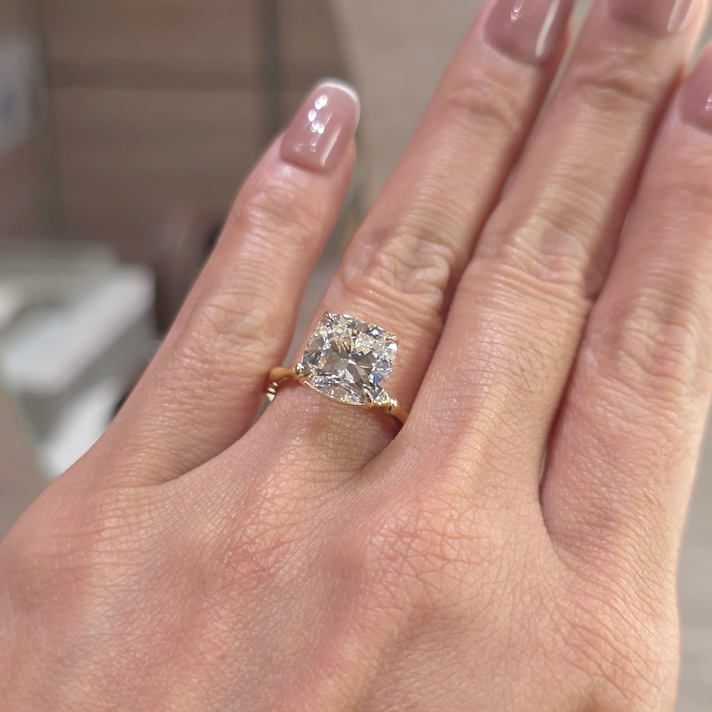 A close-up of a hand wearing a gold ring with a cushion-cut clear gemstone. The ring is shown on a finger with manicured nails, set against a softly blurred indoor background.