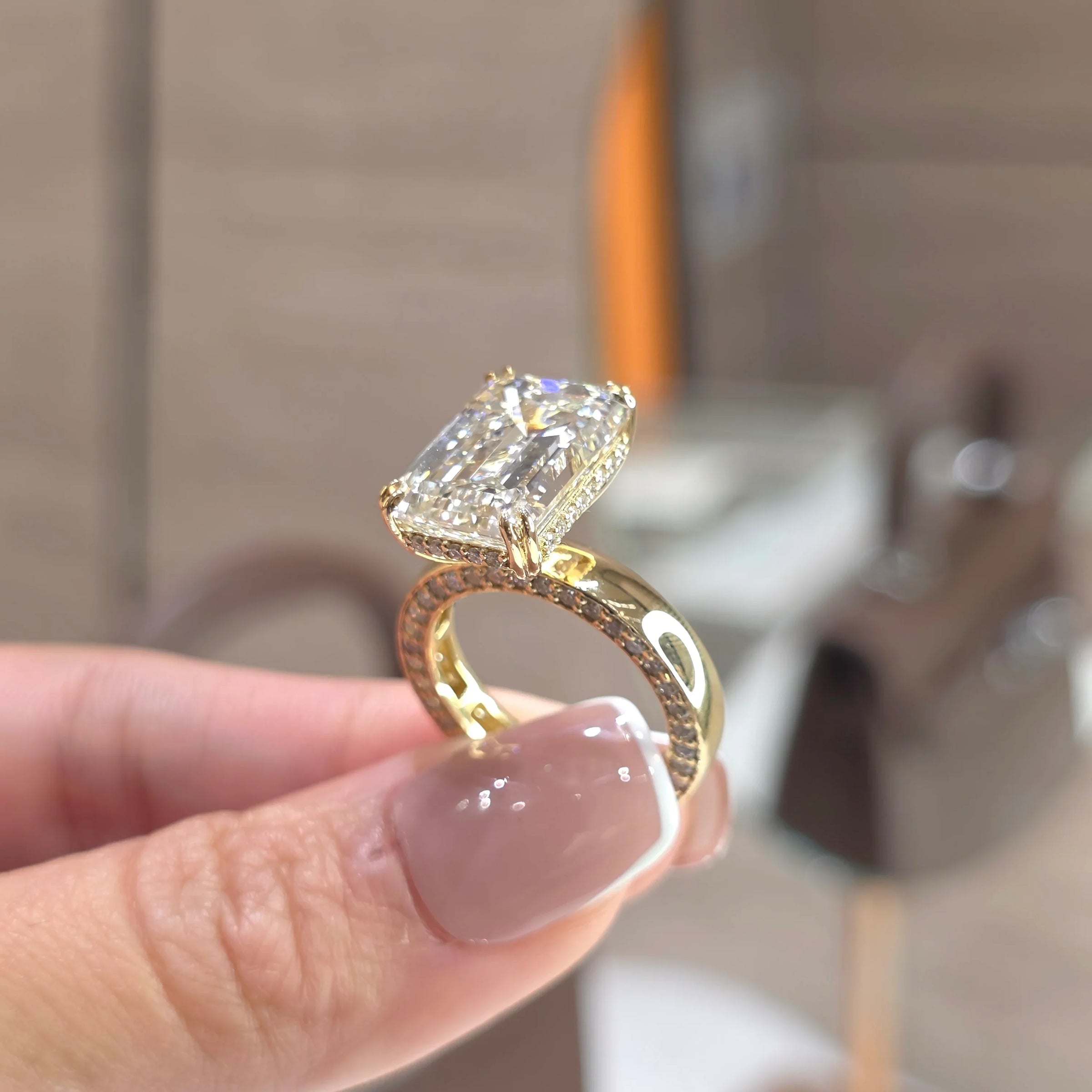 A yellow gold ring with a large rectangular clear gemstone held by a hand with a French manicure, set against a blurred indoor background.