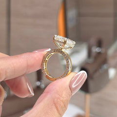 A close-up of a hand holding a gold ring with a large clear gemstone and small accent stones, set against a blurred indoor background.