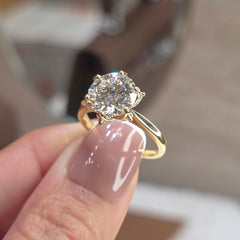 A close-up of a hand with mauve-colored nail polish holding a yellow gold solitaire engagement ring with a round brilliant-cut diamond. The background is a softly blurred indoor setting.