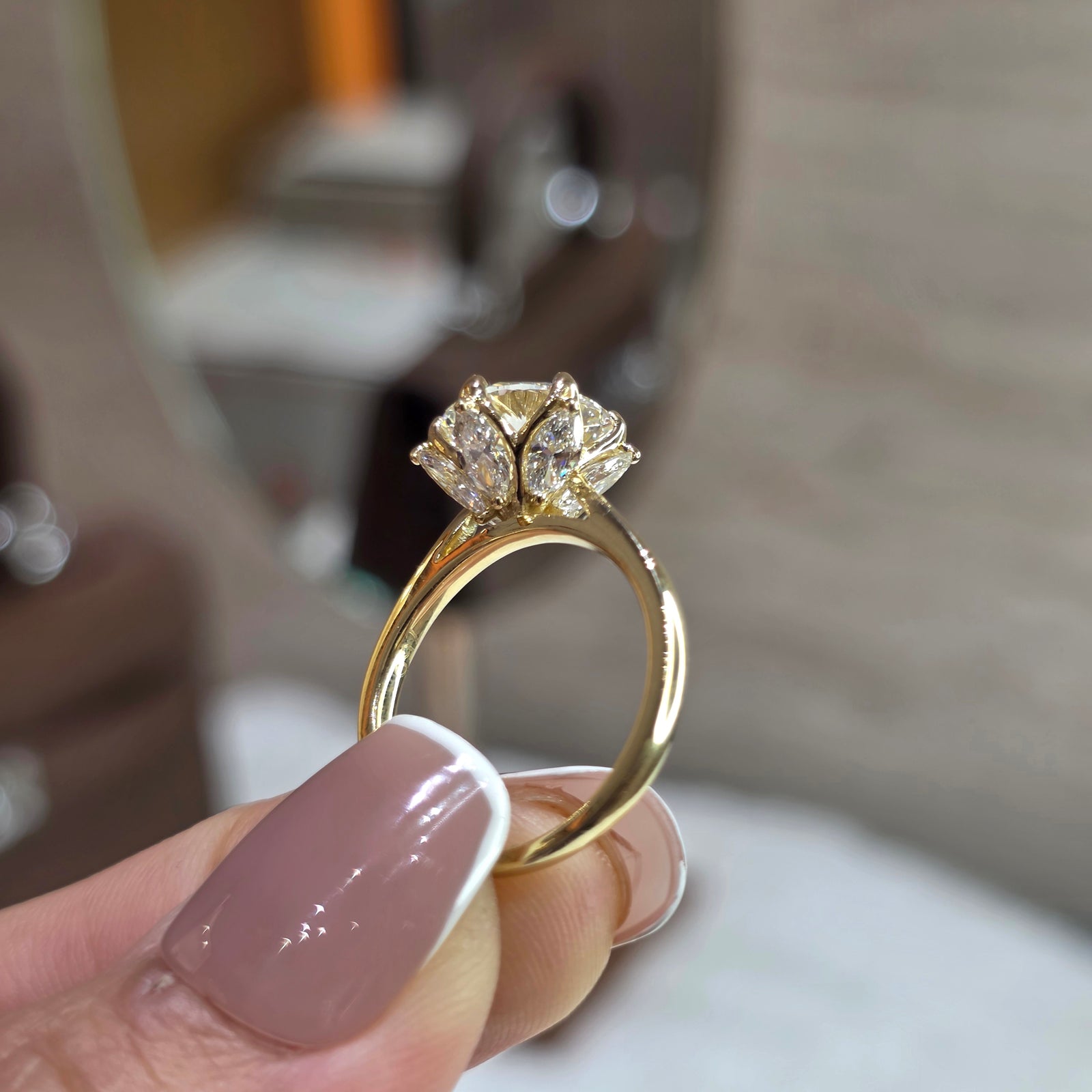 A close-up of a gold ring with a floral cluster of clear stones held by a hand with mauve French-tipped nails, with a blurred mirror in the background.