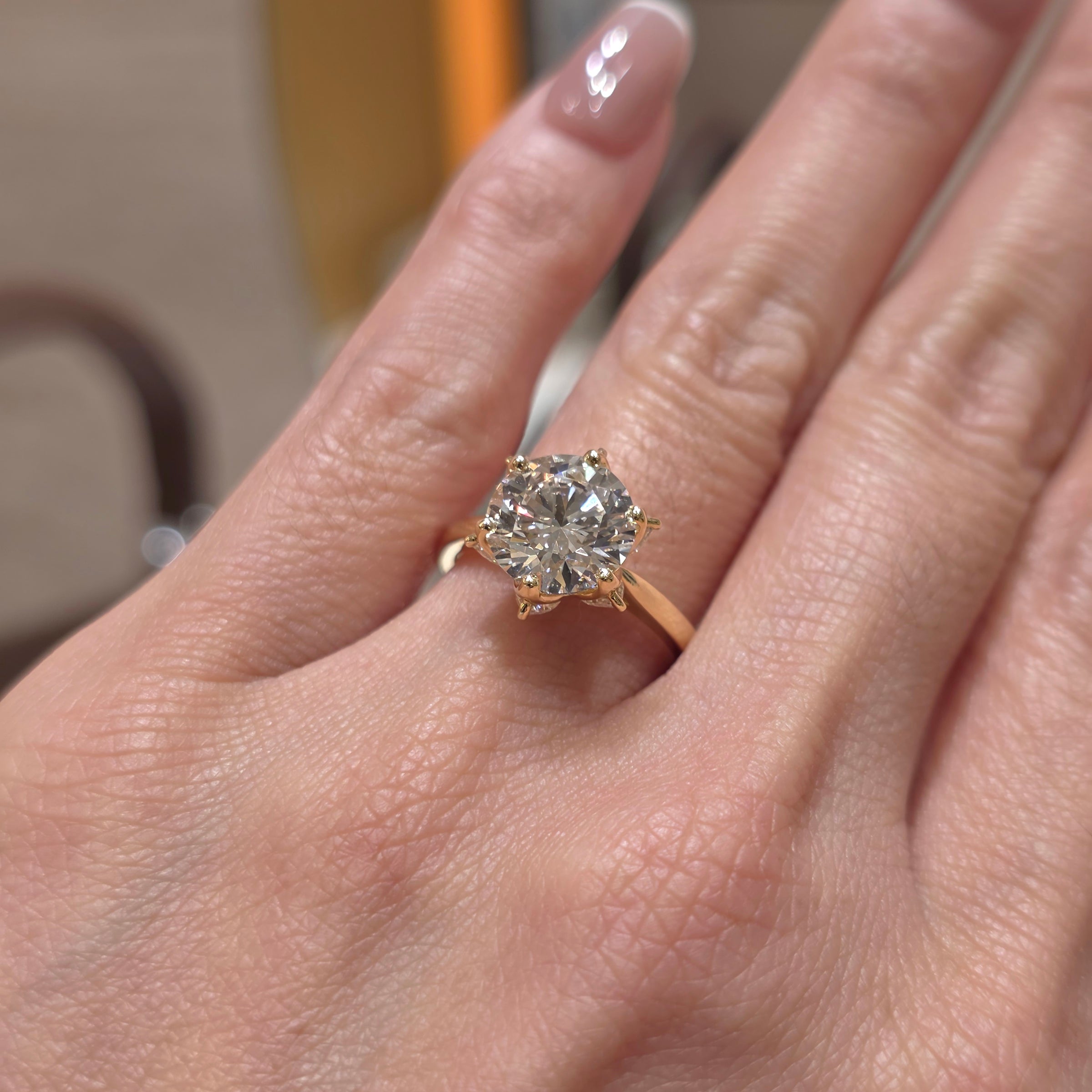 A close-up of a hand wearing a yellow gold ring with a large round brilliant-cut diamond. The ring is shown on the middle finger against a softly blurred indoor background.